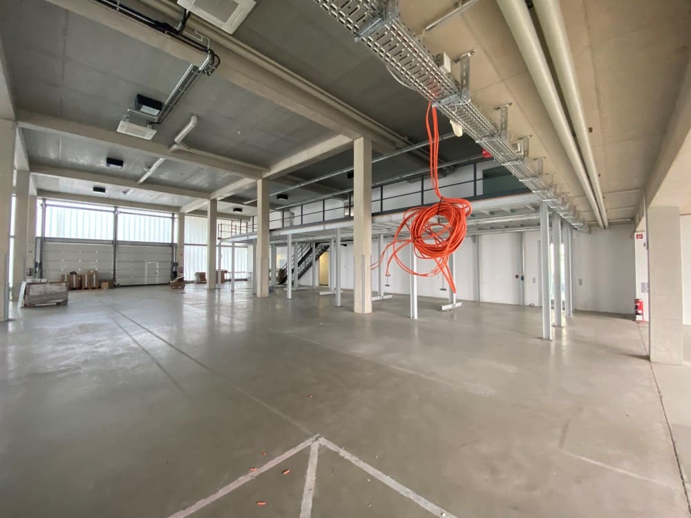 Der Innenraum einer leeren, weitläufigen Industrielagerhalle mit Betonboden, hohen Decken, metallischer Infrastruktur und roten Kabeln, die von der Decke hängen. | casaceramica