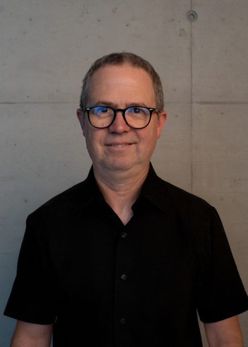 Ein Mann mittleren Alters mit kurzen grauen Haaren und Brille steht an einer schlichten Betonwand, trägt ein schwarzes Kragenhemd und lächelt leicht - perfekt für eine "Über uns"-Einführung. | casaceramica