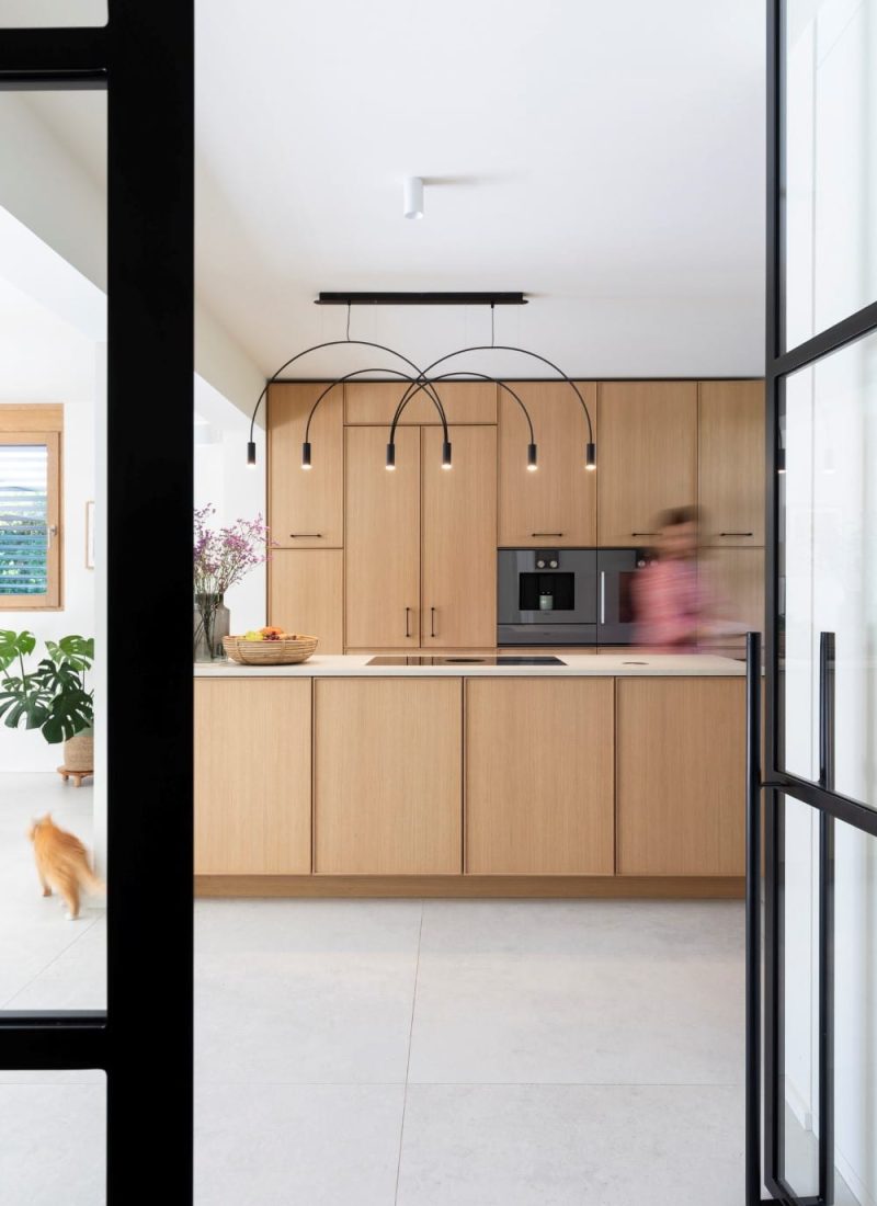 Eine moderne Küche mit hellen Holzschränken und einer Kochinsel in der Mitte sowie einer einzigartigen Deckenbeleuchtung. Im Hintergrund bewegt sich eine verschwommene Gestalt und im Vordergrund läuft eine Katze. | casaceramica